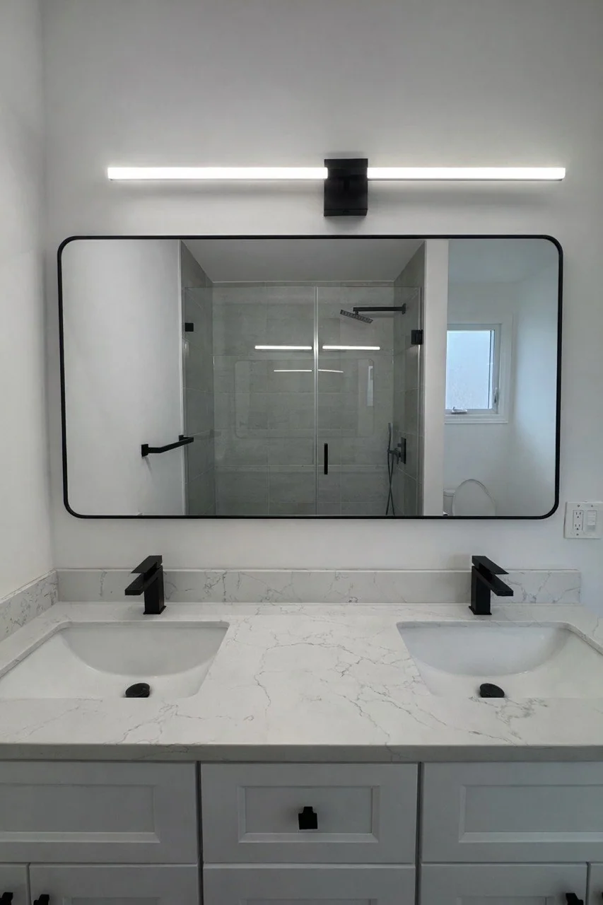 Illuminated bathroom mirror above a double sink vanity with black faucets